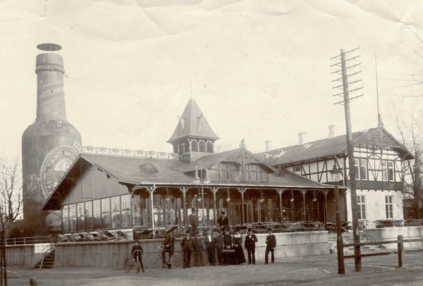 Mænd og kvinder står samlet uden for en pavillion med glasvinduer langs væggene, Bag pavillionen kan man se den store Tuborgflaske.