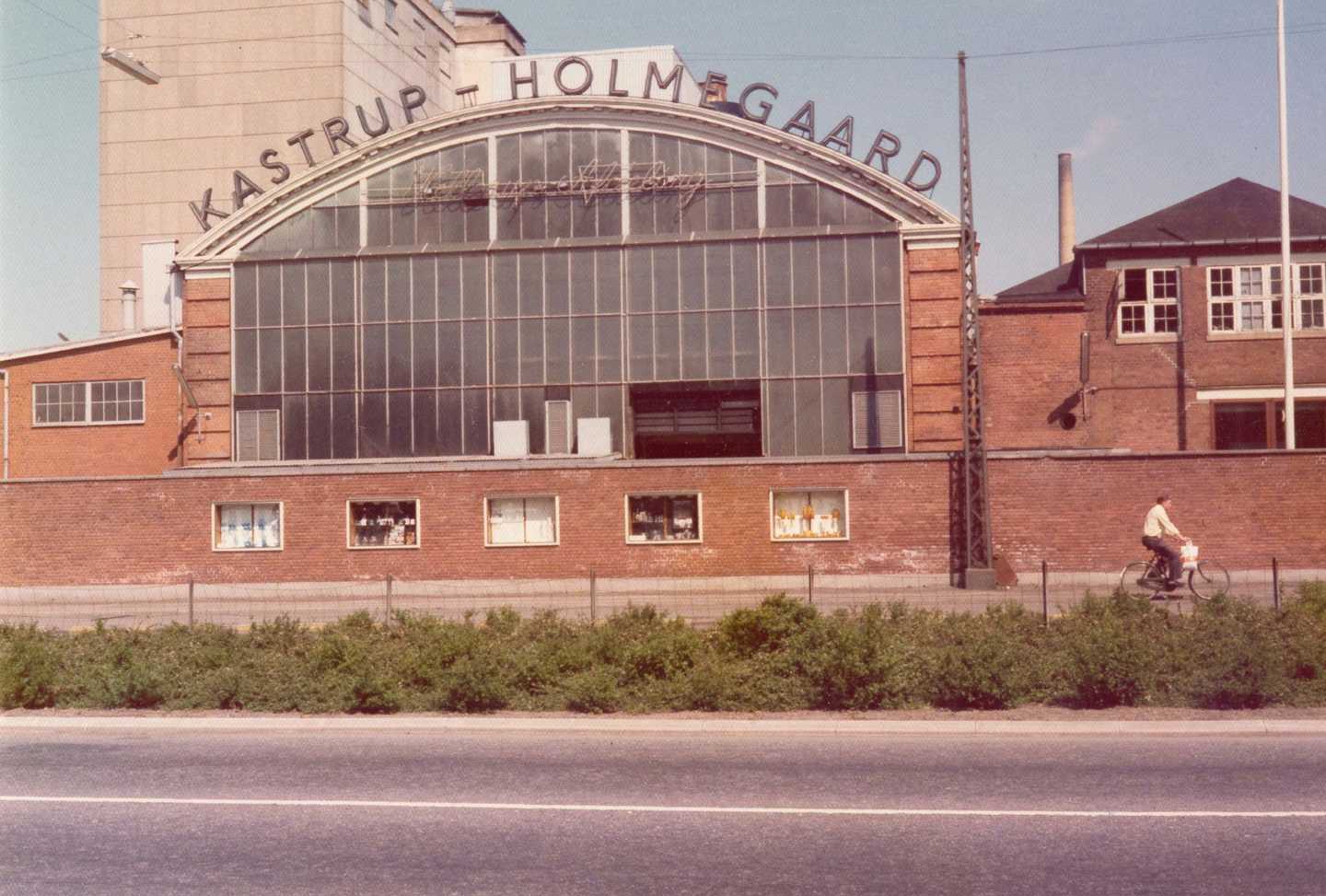 Fabriksbygning med stort glasparti og rundet tag. Langs tagkanten står "Katrup-Holmegaard" 