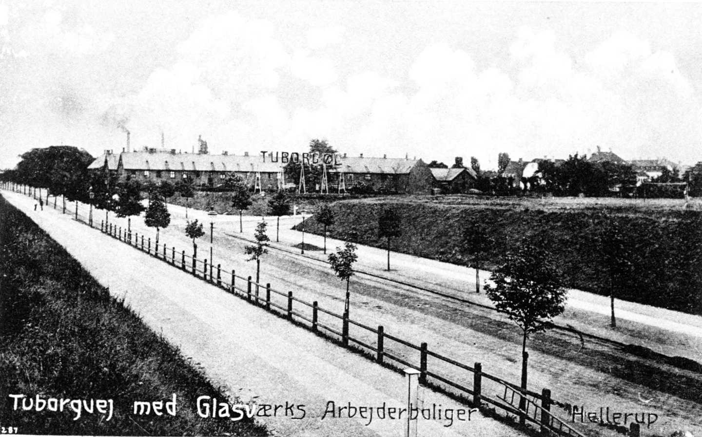 En lang lige vej med nyplantede vejtræer. I baggrunden en karre med huse, hvor der tilsyneladende er opsat et skilt på taget med teksten "Tuborg Øl".