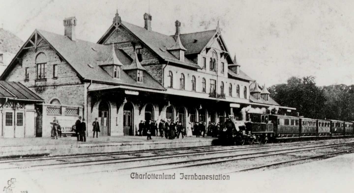 Sort/hvid foto med trykt tekst nederst i billedet - "Charlottenlund Jernbanestation". Billedet viser stationsbygningen med passagerer ventende på perronen og et damplokomotiv holdende foran.