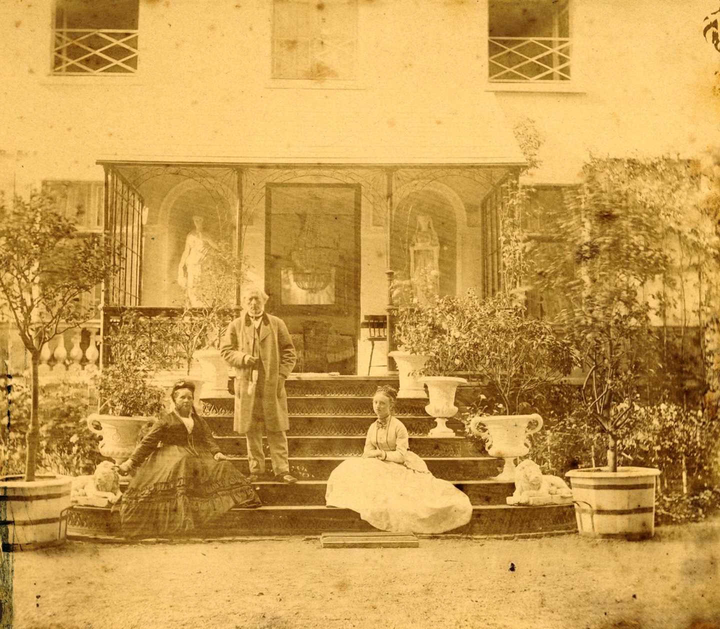Gammelt foto fra midten af 1800-tallet, to siddende kvinder i store kjoler samt en stående mand med en lang pibe på trappen ved villaen Lille Tuborg. Omkring dem står store potter med planter i og den ene kvinde hviler hånden på en løve af sten.